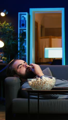 vertical video smiling man lounged on couch at home eating popcorn, watching tv