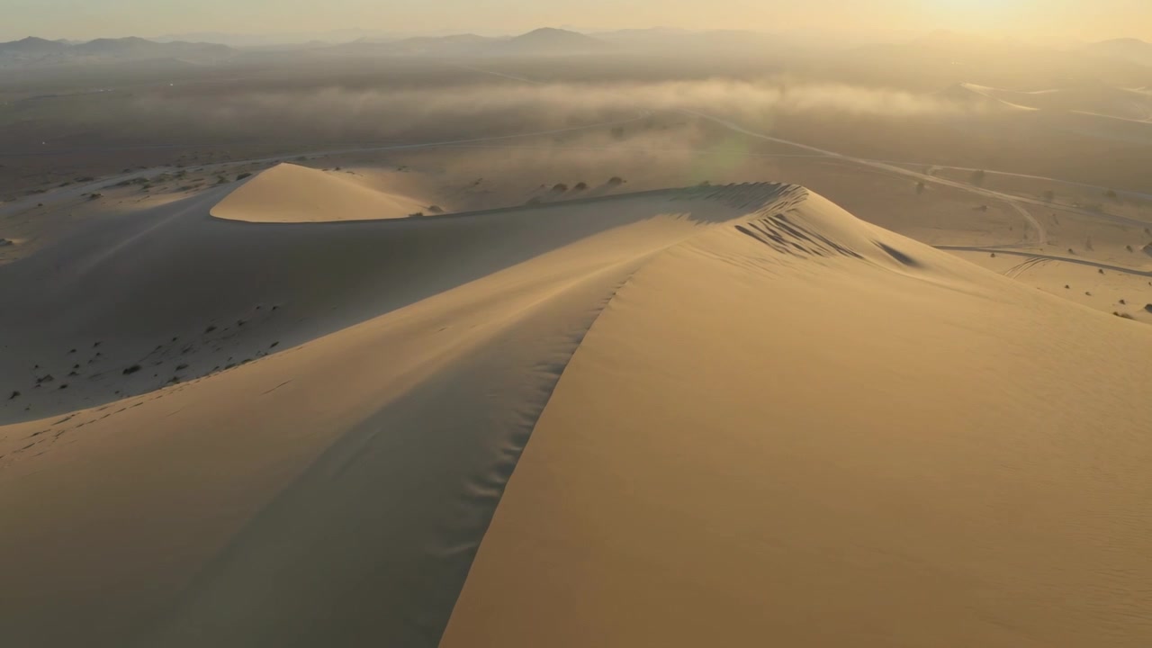 Inizio ripresa, telecamera aerea che plana lungo la cresta di una duna all'ora d'oro, rivelando increspature e una strada.