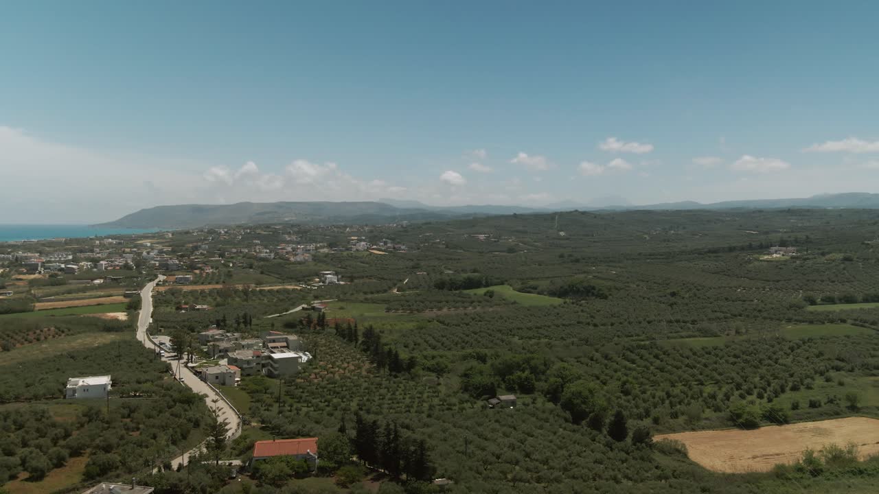 Panoramic aerial of lush olive groves and farmland stretching across the countryside near Asprouliani in Crete with hills, houses, and distant sea under clear sky