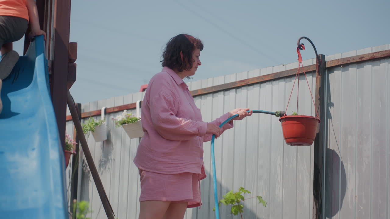 Woman Watering Plants By Backyard Pool, Casual Pink Outfit Holding Hose To Water Potted Flower Near Slide, Domestic Garden Routine Under Bright Sunlight, Relaxed Summer Chore Scene