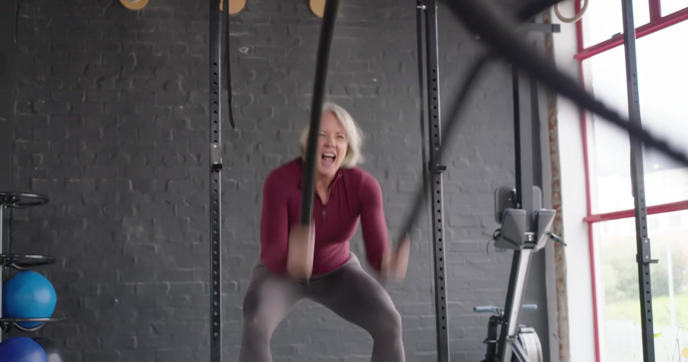 Donna anziana con top bordeaux e leggings, che afferra le battle ropes e crea onde con le corde per la forza in palestra.