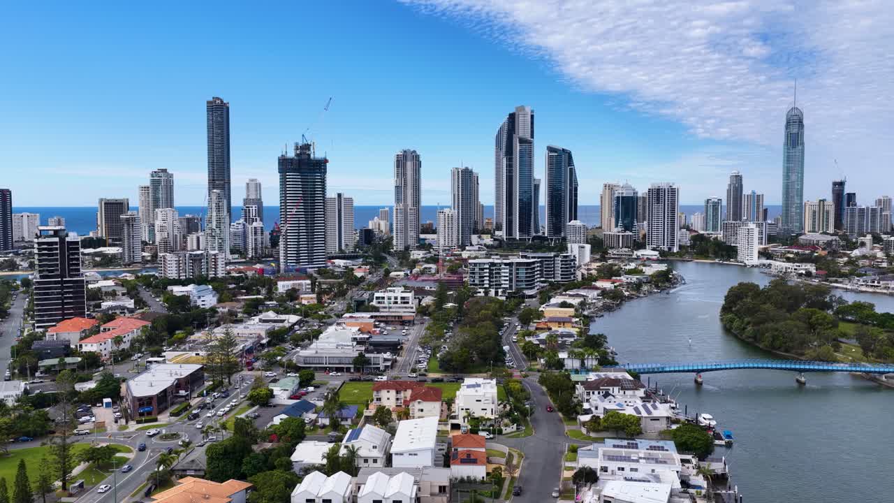 Drone camera glides above residential rooftops and waterways toward Gold Coast’s skyline, capturing high-rise buildings, clear daylight, and urban coastal scenery