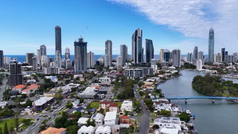 drone camera glides above residential rooftops and waterways toward gold coast’s skyline, capturing high-rise buildings, clear daylight, and urban coastal scenery