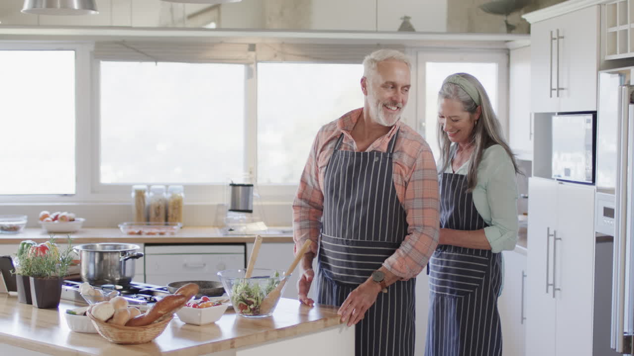 Coppia caucasica di mezza età che indossa il grembiule, cucina in cucina a casa, al rallentatore.