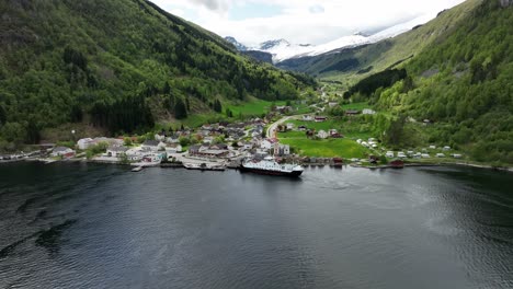 Norway-countryside-infrastructure-concept---Ferry-is-alongside-small-town-of-Eidsdal-loading-cars---Backward-ascending-aerial-during-spring-with-green-hillsides-and-snow-capped-mountain-background