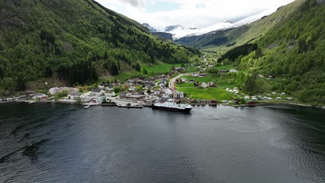 norway countryside infrastructure concept - ferry is alongside small town of eidsdal loading cars - backward ascending aerial during spring with green hillsides and snow capped mountain background