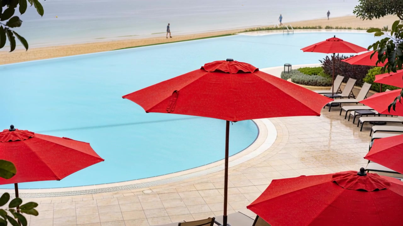 Inquadrando ombrelloni rossi con una piscina blu sul ponte di un resort, due persone camminano entrando da sinistra lungo la spiaggia.