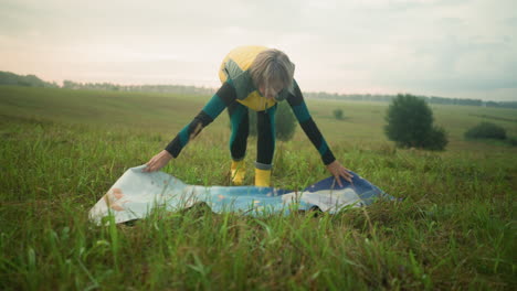 frau in gelber jacke und stiefel breitet ihre matte auf einem grasbewachsenen feld aus und bereitet sich auf eine outdoor-aktivität in einem weiten offenen feld mit bäumen in der ferne und bewölkten himmeln über dem kopf vor