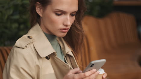 caucasian female student using smartphone and drinking coffee outdoors.