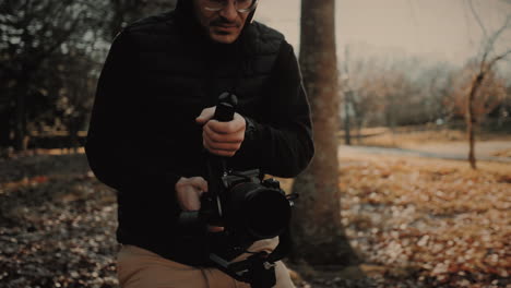 feliz y alegre joven camarógrafo caucásico con gafas, un chaleco negro y una capucha, usando una cámara de video y un estabilizador para grabar la temporada de otoño en un bosque en la naturaleza