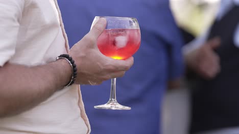 person holding glass of red cocktail with ice cubes at an outdoor gathering