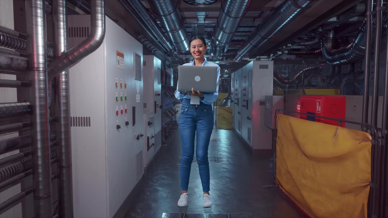 Full Body Of An Asian Female Professional Worker Standing With Her Laptop In Engine Control Room, She Is Looking At The Camera With A Smile