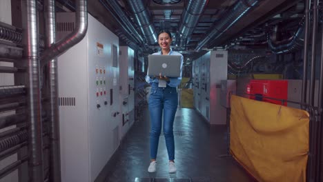 full body of an asian female professional worker standing with her laptop in engine control room, she is looking at the camera with a smile