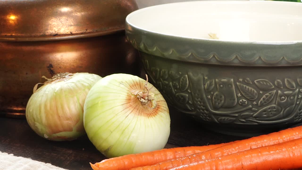 Close up of picking up a yellow or Spanish onion off a wooden cutting board in the kitchen. Making Italian Wedding Soup for dinner. Carrots, onions, large green ceramic bowl and vintage tea kettle.