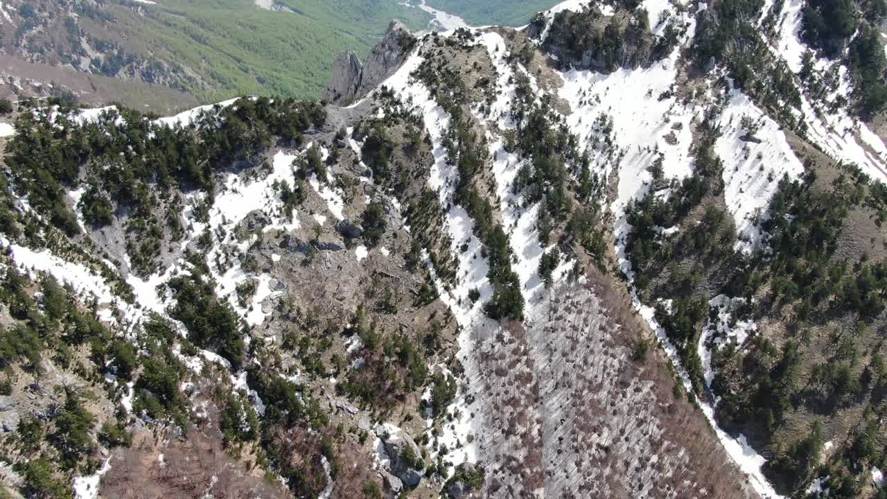 una vista de avión no tripulado en albania volando en los alpes que muestra un pico de montaña nevado y rocoso entre dos verdes valles forestales en el