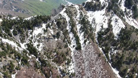 Drone-view-in-Albania-flying-in-the-alps-showing-a-snowy-and-rocky-mountain-peak-between-two-green-forest-valleys-in-Theth