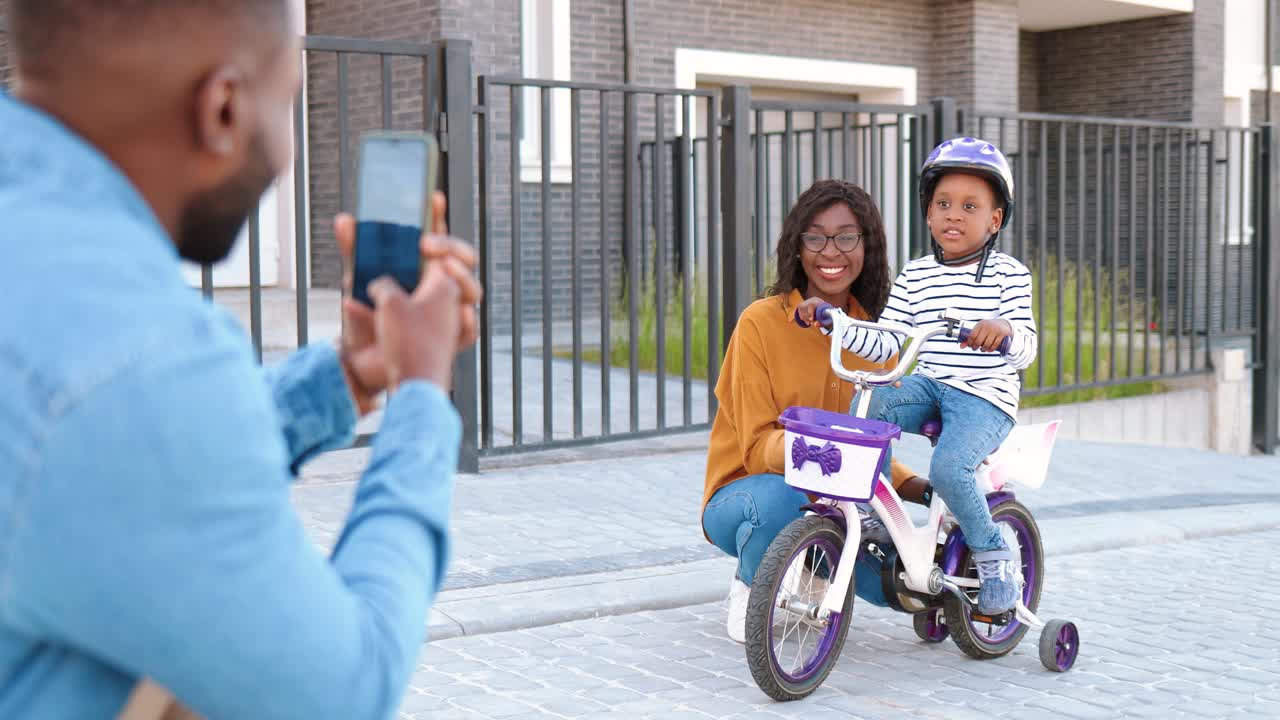 Back view on African American father taking photo with smartphone camera of wife and daughter on bike