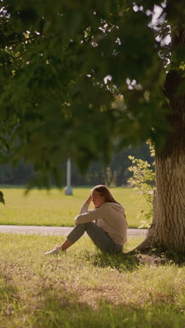 woman sits on grassy ground under shaded tree, thoughtfully resting hand on her leg, surrounded by greenery, background includes park setting with poles and open field