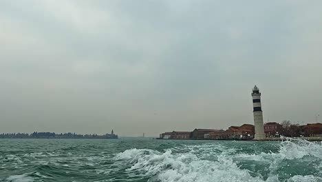 Low-angle-water-surface-view-of-Murano-lighthouse-seen-from-sailing-motorboat,-Italy