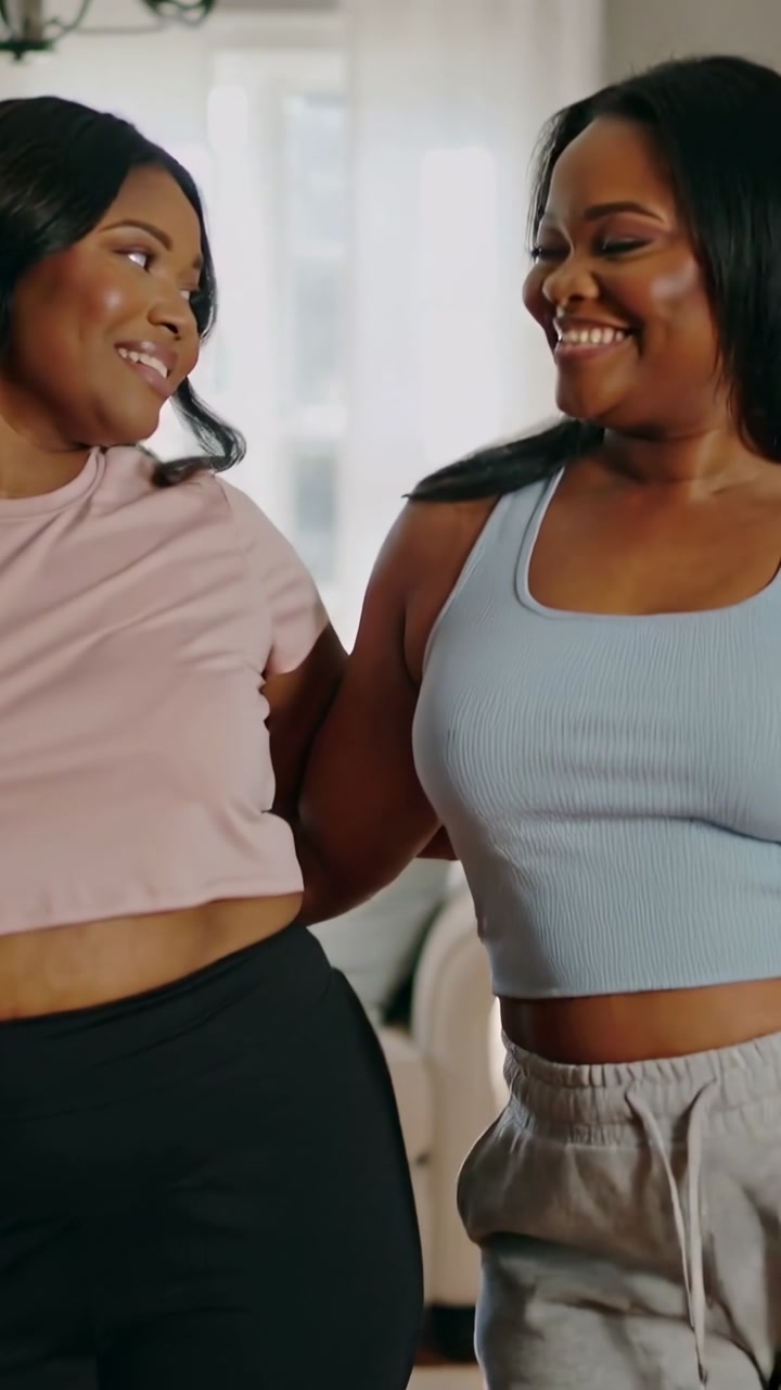 Vertical video: Linking arms, two women sharing smiles at home by couch in crop tops showing warmth