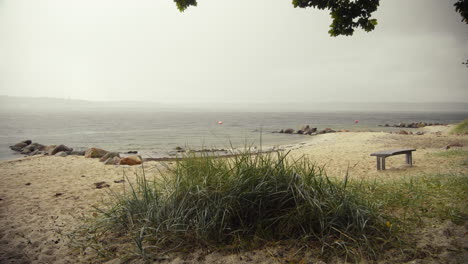 Leerer-Strand-Mit-Dünengras-Und-Holzbank-Vorne-An-Einem-Bewölkten-Tag-Am-Fjord