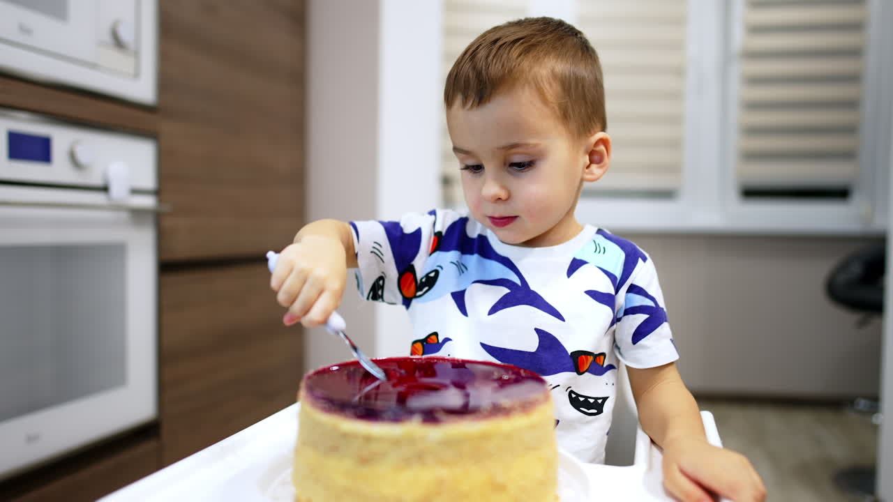 Lovely toddler boy puts the fork into a big cake in front of him. Parent's hand shows number three candle burning and kid blows it.
