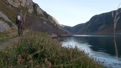 Ein-Mann-Wandert-Von-Einer-Kamera-Im-Schottischen-Hochland-Mit-Bergklippen-Und-Einem-Meeresloch-Im-Hintergrund,-Während-Im-Vordergrund-Heidekraut-Und-Gras-Sanft-Im-Wind-Wehen