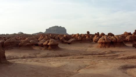 Gorgeous-dolly-in-aerial-drone-shot-of-the-beautiful-Goblin-Valley-Utah-State-Park-approaching-strange-mushroom-rock-formations-with-a-large-red-Butte-in-the-background-on-a-warm-sunny-summer-day