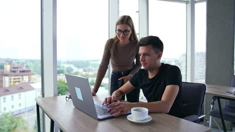 colleagues discussing work looking at computer screen. female teammate makes suggestions pointing at the laptop. big light office indoor backdrop.