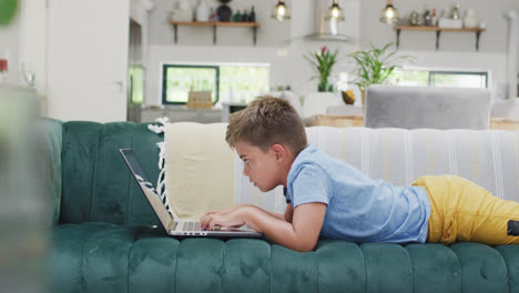 Feliz-Niño-Caucásico-Tumbado-En-El-Sofá-Y-Usando-Una-Laptop,-Cámara-Lenta