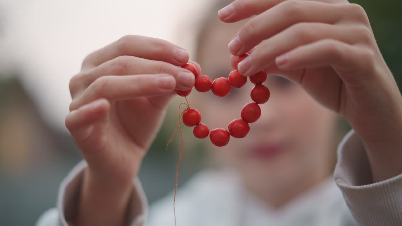 Mani che formano un cuore con perline rosse, ragazza all'aperto in primo piano. Dita delicate che creano un cuore con perline rotonde su un filo sottile, sfondo bokeh morbido, luce calda del crepuscolo, atmosfera nostalgica del fatto a mano.