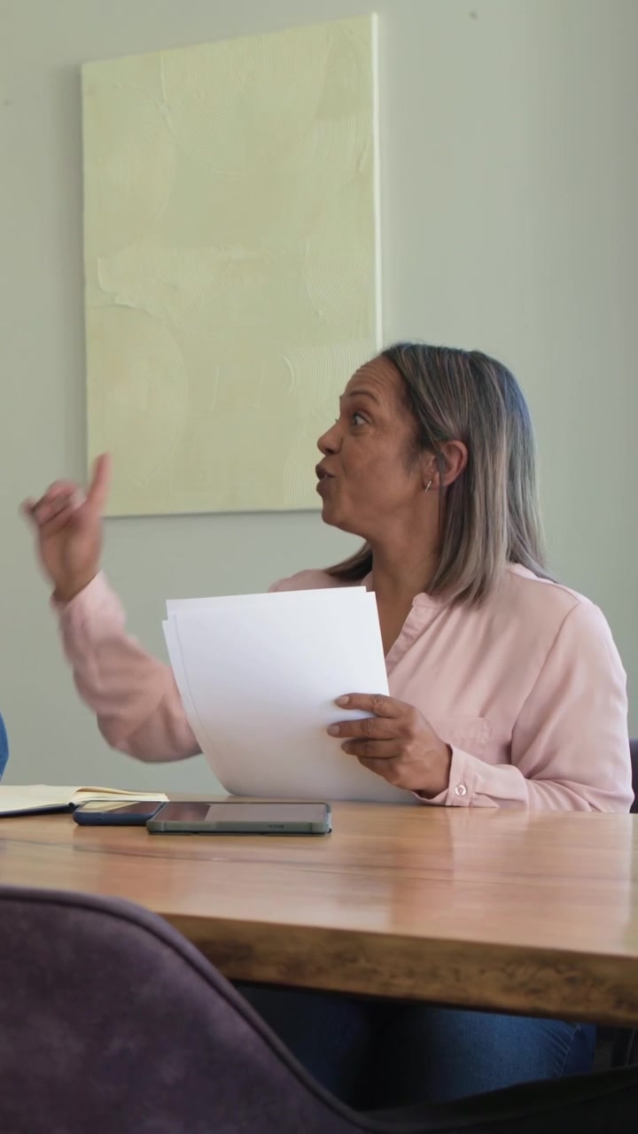 Video verticale: Riunione che inizia, donna con maglietta rosa che legge documenti a un tavolo di legno, toccando un tablet