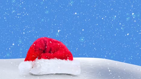 Digitale-Animation-Von-Schnee,-Der-über-Die-Weihnachtsmütze-Auf-Schnee-Vor-Blauem-Hintergrund-Fällt