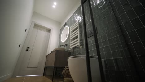 un baño contemporáneo con azulejos de color verde oscuro, un espejo oval con iluminación de fondo, un radiador blanco y una vanidad de madera, que emite un ambiente moderno y funcional