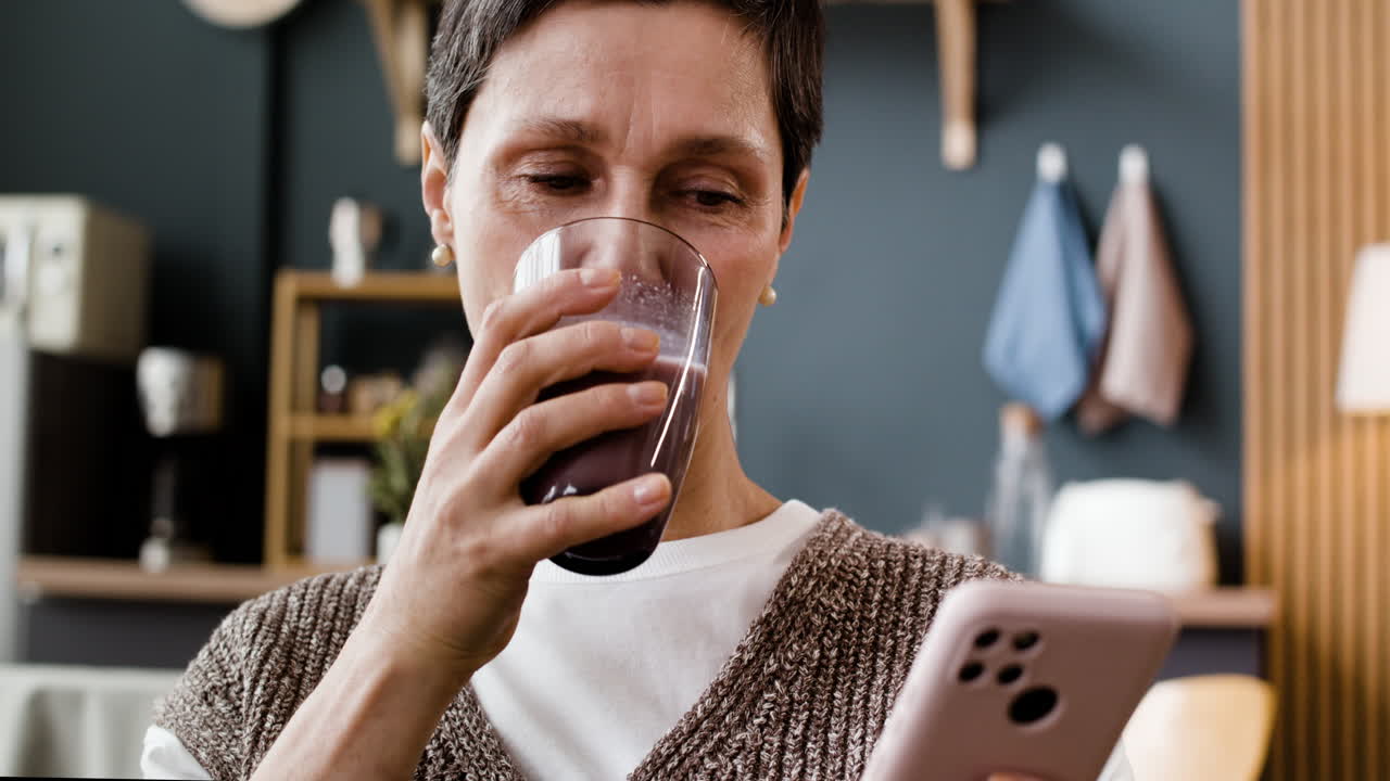 A woman drinks from a glass while using her smartphone.