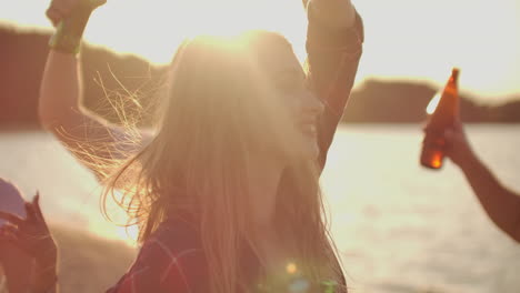 una joven feliz está bailando en la fiesta al aire libre con sus amigas en la playa. mujer de pelo rubio largo está volando en el viento. sonríe y disfruta de la puesta de sol en la costa del lago con cerveza.