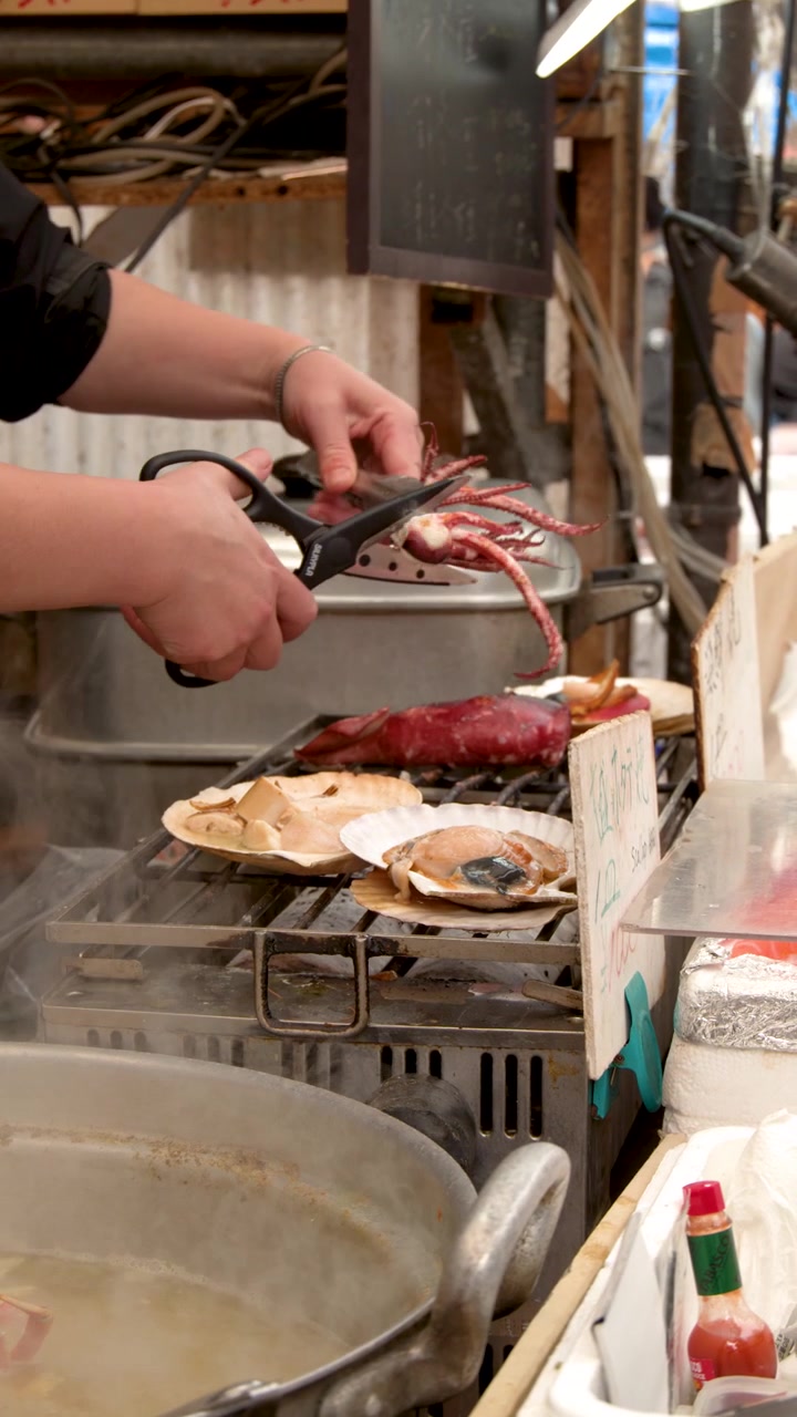 Un venditore di cibo da strada usa pinze e forbici per preparare capesante fresche su una griglia.