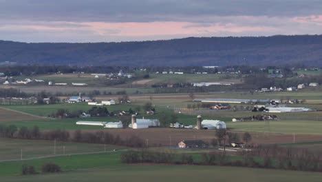 Aerial wide shot showing idyllic farm fields and farm houses in american countryside. Cloudy day behind forest mountains. barn with storage and green fields. Driving cars on rural road.