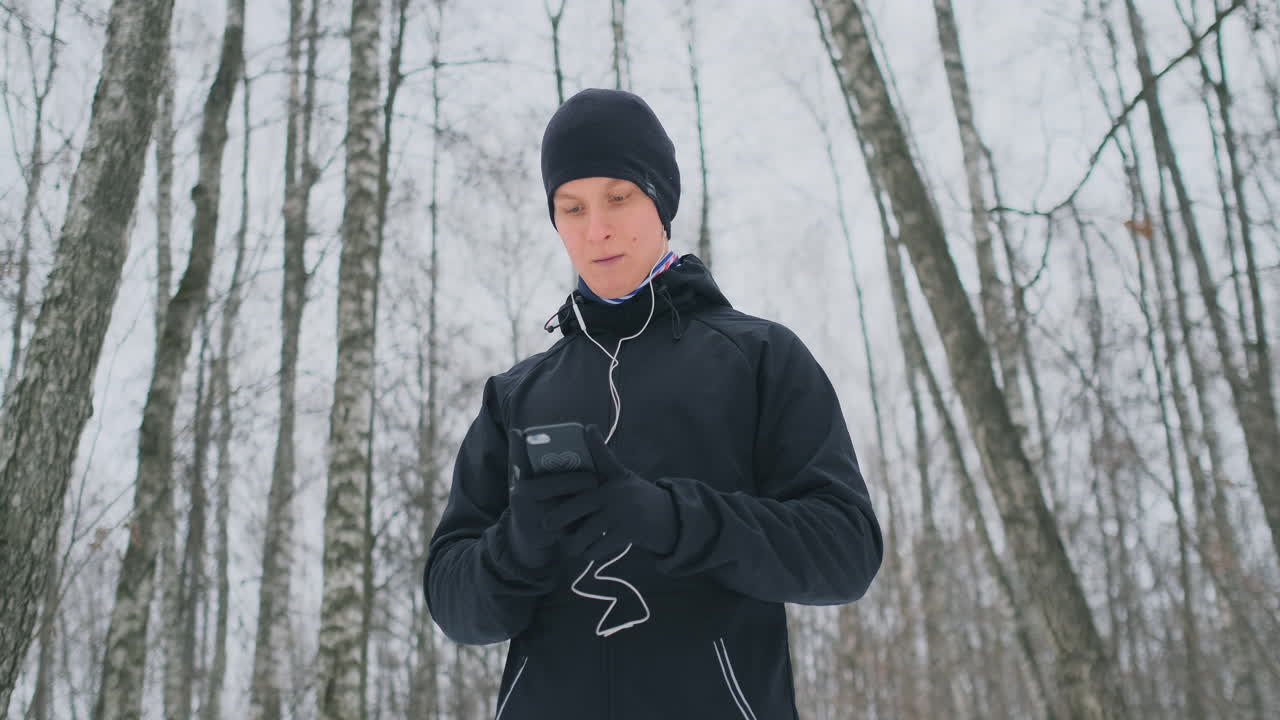 겨울에 검은 방아쇠를 입은 청년이 공원에서 조깅을 준비하고 있으며 스마트폰으로 음악을 틀고 음성 보조 장치와 함께 숲속을 뛰어다니면서 사업을 하고 건강한 생활 방식을 선도하고 있습니다.
