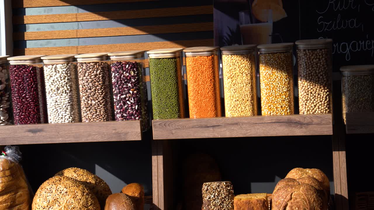 Morning sun shining across plastic bins of legumes, grains, and fresh bread displayed in market.