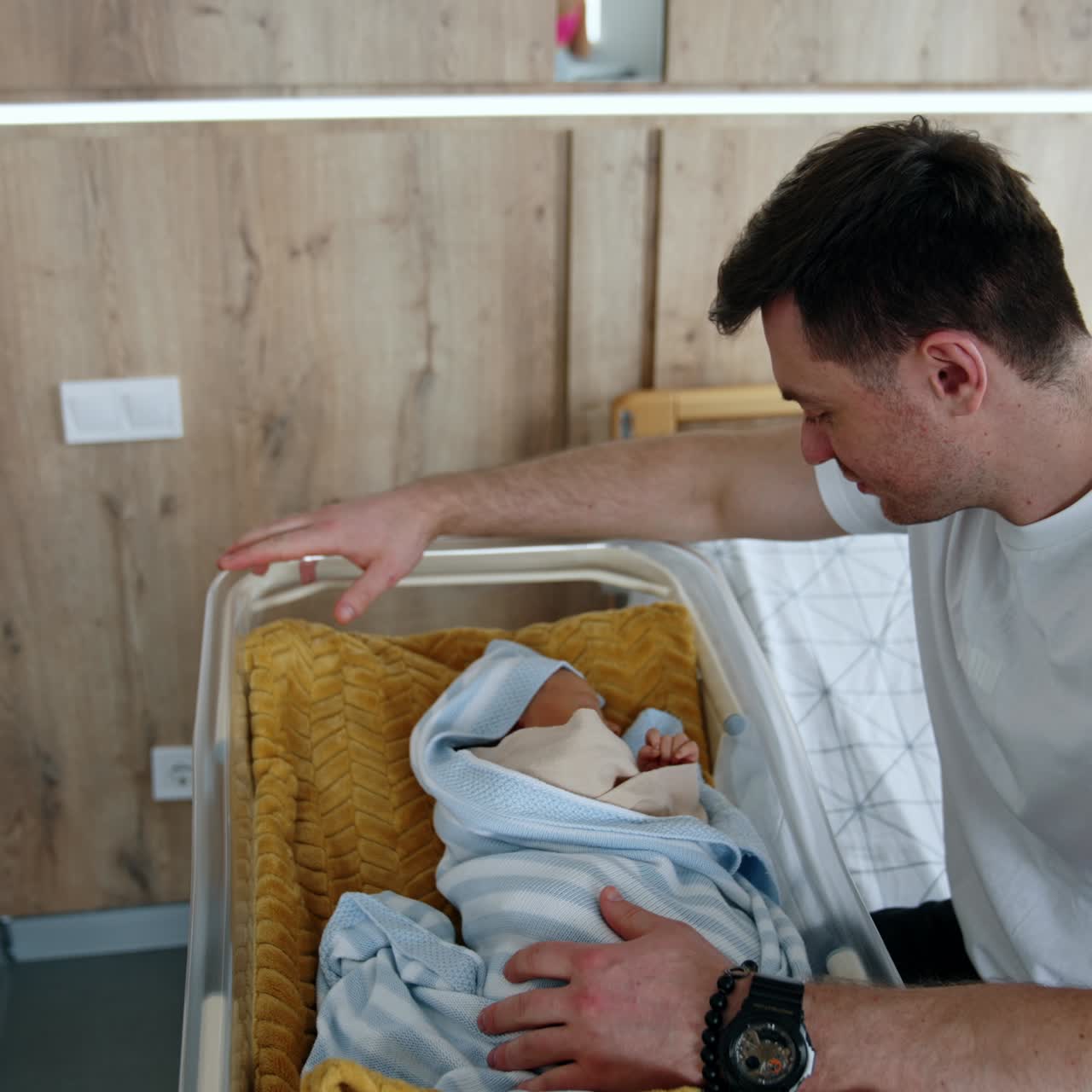 Happy dad sits at the crib with his newborn child. Caring father checks the blankets and talks to his baby