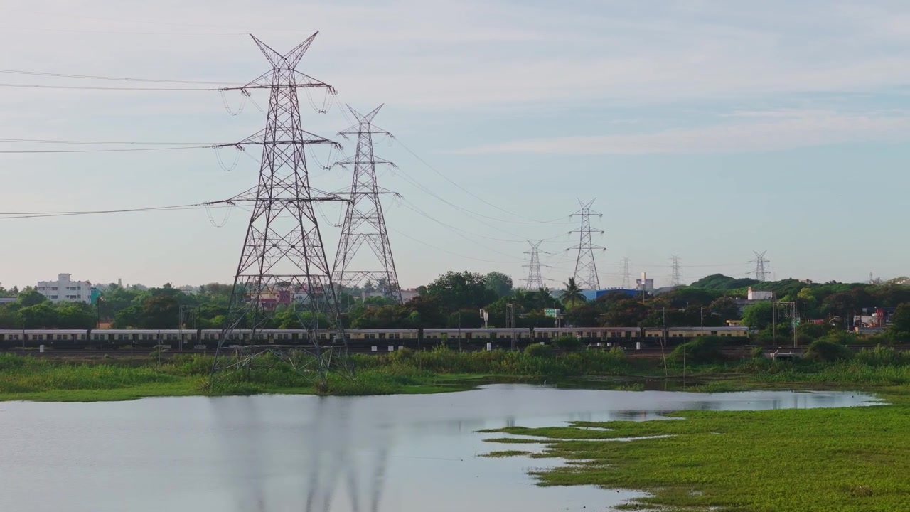 Train passing beside a lake featuring power transmission towers, overhead power lines, calm water, green shoreline, and urban landscape near Chennai, India