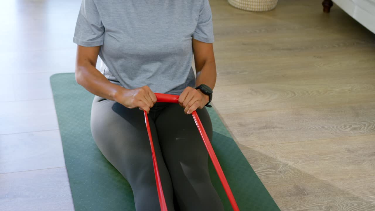Donna afroamericana in abbigliamento sportivo che tiene una fascia elastica, tirandola verso il petto su un tappetino da casa per l'allenamento della forza