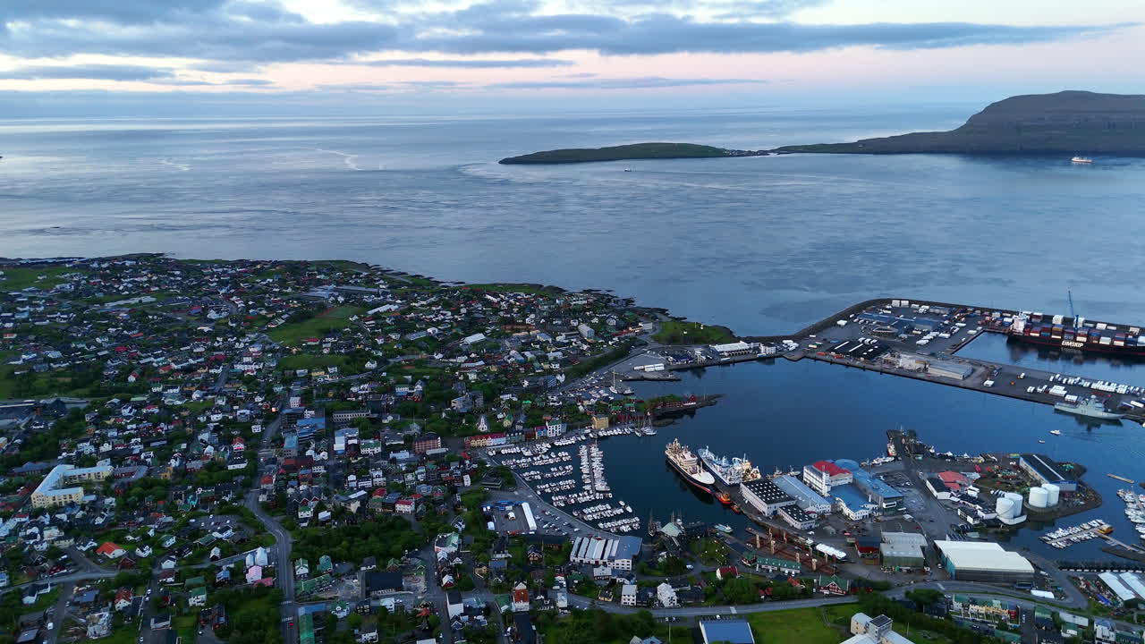 Cinematic aerial view of colorful village houses along a fjord in the Faroe Islands, surrounded by dramatic green mountains, black sand beach, and misty coastal scenery