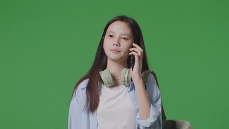 close up of asian teen girl student with a backpack talking on smartphone while walking in the green screen background studio