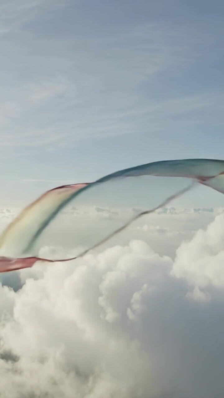 Vertical video: Entering from right, sheer rainbow ribbon undulating above clouds, driven by breeze