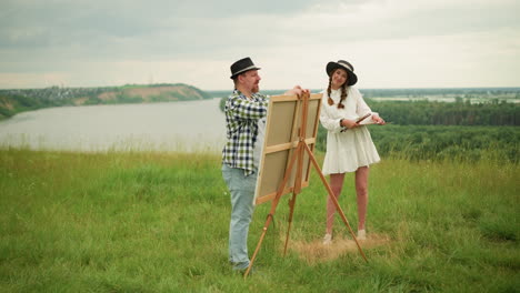 two partners are seen in a grassy field. the man, wearing a black hat and plaid shirt, is setting up a drawing board on a wooden tripod. nearby, a woman in a white dress stands smiling as she watches