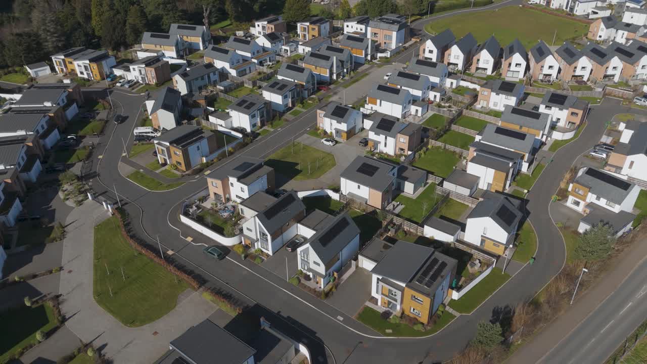 Aerial glide along curving streets of low-rise solar-ready homes with private plots, hedges and roundabouts showcasing balanced suburban development