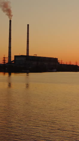 evening river on power plant background. smoke from industrial pipes goes into the air against the setting orange sun. silhouette of a factory near the lake. vertical video