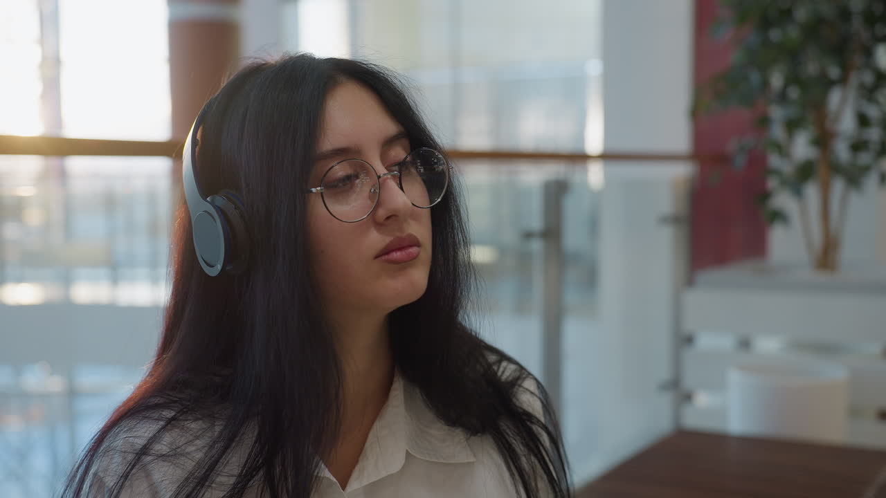 Young woman with glasses and headphones gazes thoughtfully in modern indoor setting before looking down, surrounded by soft daylight and glass panels, reflecting calm and introspective mood